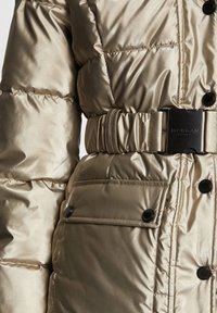 Veste en métal doré matelassée avec une taille ceinturée, présentant une texture lisse, des boutons noirs et une poche plaquée.