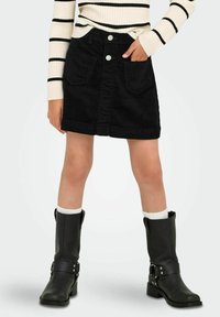 Bambina che indossa una gonna di velluto a coste nera con tasche frontali, un maglione bianco e nero a righe, calze bianche e stivali neri fino al polpaccio, in piedi con una mano in tasca.