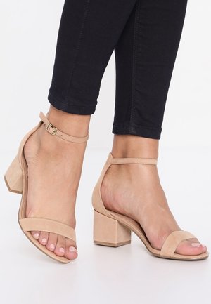 Pieds portant des sandales à talons blocs nude avec des brides à la cheville, associées à un pantalon noir coupé au-dessus de la cheville sur un fond blanc.
