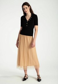 Schwarzes, geripptes Kurzarmoberteil mit goldenen Knöpfen, kombiniert mit einem beigen, plissierten Tüllrock. Glänzende, schwarze spitze Ballerinas runden das Outfit ab.