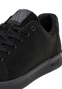 Gros plan sur une basket basse noire avec des lacets noirs, un détail perforé sur le côté, un col rembourré et une semelle en caoutchouc texturée avec le nom de la marque sur le talon.