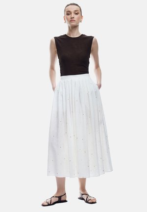 Brun ærmeløs top, hvid lang nederdel med plisseringer og subtil blomsterdesign, parret med sorte snøresandaler.