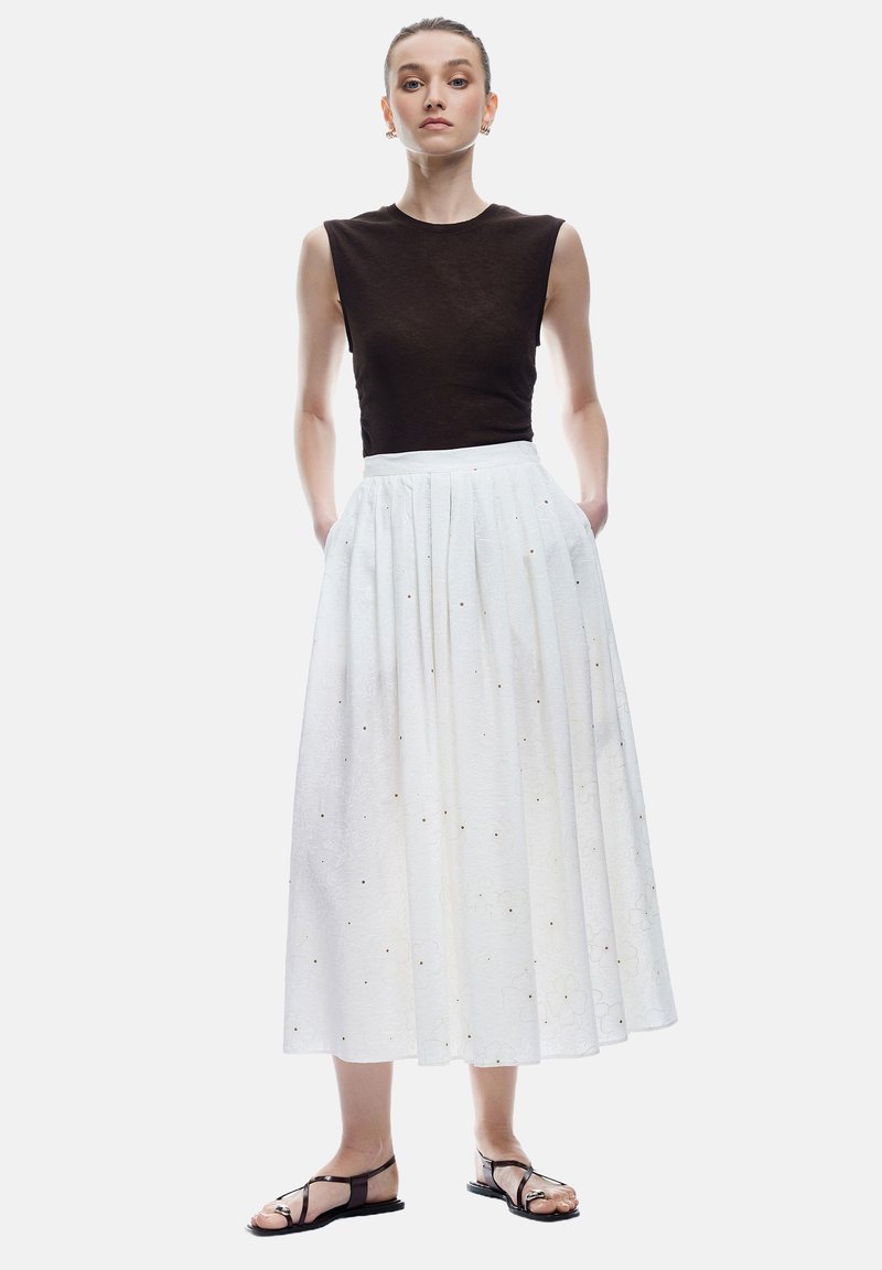 Top sans manches marron, jupe longue blanche avec des plis et un motif floral subtil, associée à des sandales noires à lanières.
