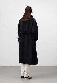 Schwarzer Wollmischmantel mit tailliertem Gürtel, übergroßem Kragen und langen Ärmeln, kombiniert mit weißen Hosen und schwarzen High Heels.