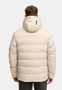 Doudoune beige avec capuche, présentant des sections matelassées horizontales et des poignets élastiques. Comprend une petite étiquette de marque au dos de la capuche.