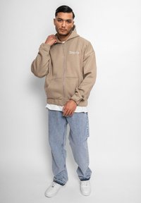 Beige zip-up hættetrøje med en frontlomme, parret med lyseblå jeans og hvide sneakers. Hættetrøjen har et lille logo på brystet.