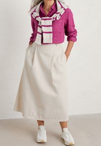 Kvinna i lila skjorta med knappar, randig tröja över axlarna, beige midiklänning och vita sneakers står mot en vit vägg.