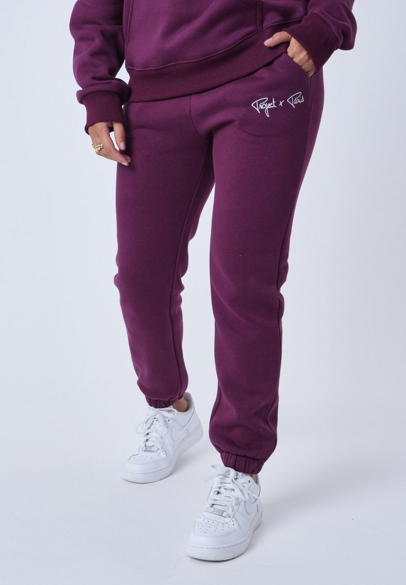 Pantalones de chándal burdeos con puños elásticos, bolsillos laterales y detalles de logo en blanco, combinados con zapatillas blancas. Material suave y ajuste relajado.