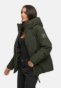 Woman wearing an unzipped dark green puffer jacket over a black top and black jeans with one hand in pocket, facing left on plain background.