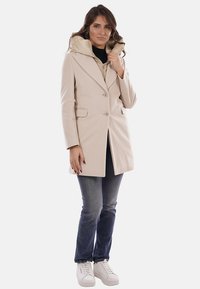 Cappotto beige doppiopetto con cappuccio, caratterizzato da una texture liscia, tasche frontali e una vestibilità su misura, indossato sopra jeans scuri e sneakers bianche.