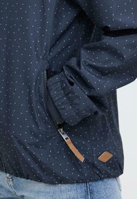 Veste bleu marine à petits pois blancs, avec poignets élastiques, poche zippée à l'avant et tirette en cuir marron, portée sur un jean bleu.