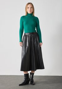 Grønn ribbestrikket turtleneck-genser sammen med svart folderleilig lærskjørt og svarte ankelstøvletter, satt mot en lys bakgrunn.