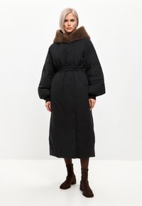 Sort puffjakke med en brun faux fur hætte, cinchet talje og lange ærmer. Jakken strækker sig til midt på læggen, har en afslappet pasform og frontlommer.