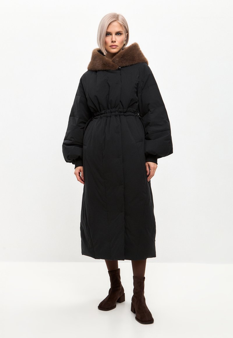 Sort puffjakke med en brun faux fur hætte, cinchet talje og lange ærmer. Jakken strækker sig til midt på læggen, har en afslappet pasform og frontlommer.
