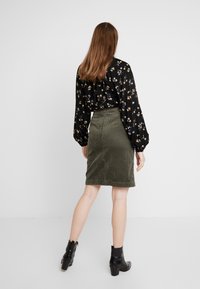 Blouse noire à fleurs à manches longues avec des manches amples, associée à une jupe crayon en velours côtelé vert olive et des bottines noires à talons.