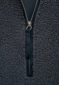 Pullover in pile nero con una cerniera in argento visibile, con una superficie testurizzata e un tirante in camoscio. La vista ravvicinata enfatizza i dettagli.