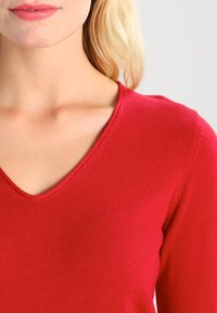 Czerwony sweter w serek o teksturze w prążki. Zbliżenie na dekolt i ramiona, z gładkimi szwami i dopasowanym krojem.