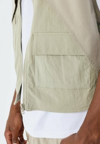 Gilet color khaki chiaro con una finitura testurizzata, dotato di una cerniera frontale, due tasche a toppa e un design a strati. Maglietta bianca visibile sotto.