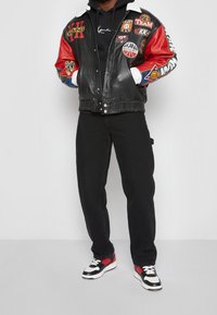 Czarna skórzana kurtka bomber z czerwonymi i niebieskimi rękawami, ozdobiona wieloma kolorowymi logo. Czarne spodnie i czarne buty sportowe z czerwonymi akcentami.