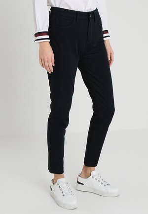 Navy venyttävät housut slim-fit-mallilla, joissa on sivutaskut ja vetoketju. Yhdistettyinä valkoisiin matalapohjaisiin tennareihin ja valkoiseen paitaan, jossa on raidallisia yksityiskohtia.