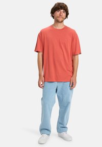Man staat, gekleed in een losse rode T-shirt, lichtblauwe jeans en witte sneakers tegen een effen witte achtergrond.
