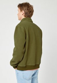 Olijfgroene fleece jas met hoge kraag, gestructureerd oppervlak en geribbelde manchetten. Het ontwerp heeft een ontspannen pasvorm en een elastische zoom.