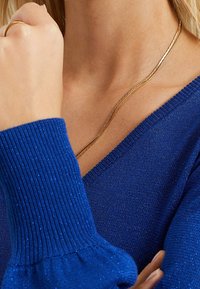 Blå stickad tröja med ribbade ärmar. Har en v-ringad design. Ett gyllene halsband vilar mot huden, vilket ger kontrast.