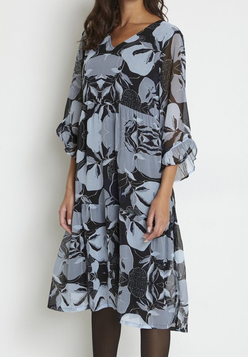 Femme portant une robe noire longueur genoux avec de grands motifs floraux gris clair et des manches trois-quarts transparentes, debout sur un fond blanc.