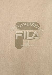 Il tessuto beige presenta un logo ricamato e testurizzato con la scritta "Established Fila" e "In 1911," in una sfumatura di verde chiaro contrastante.
