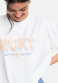Biele tričko s krátkym rukávom so veľkým grafickým prvkom "ROXY" v oranžovej a modrej farbe, pod ktorým je modrý nápis "ACTIVE BY NATURE".