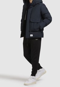 Granatowa kurtka puchowa z kapturem i przednimi kieszeniami, zestawiona z czarnymi joggerami z logo oraz białymi wysokimi trampkami.