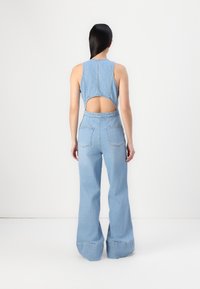 Combinaison en denim bleu clair avec un design sans manches, découpes au dos et pantalon large, dotée de deux poches arrière et de coutures visibles.