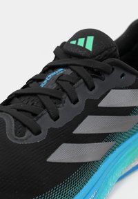 Chaussure de course Adidas noire avec des rayures grises, une semelle bleue et le texte "Supernova Rise 2" près des lacets noirs sur un fond blanc.