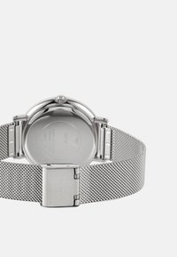 Reloj de acero inoxidable plateado con una correa de malla, caja redonda y cierre de broche. La parte trasera presenta una superficie lisa con detalles grabados.