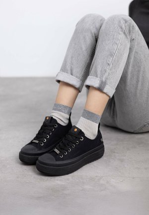 Zapatillas negras de lona con un acabado texturizado, puntera redonda y suela de goma. Combinadas con calcetines grises con acentos plateados y jeans grises remangados.