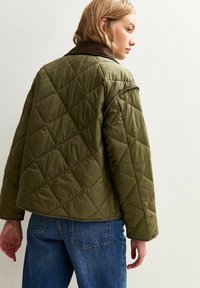 Vrouw in een groen gewatteerd jack met een donkere kraag en blauwe jeans, zijwaarts gekeerd tegen een effen lichte achtergrond.