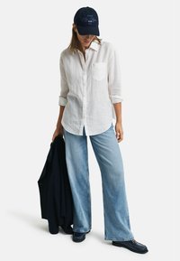 Witte linnen overhemd met knopen, lichtblauwe wijde jeans, navy pet met geborduurde tekst, houdt een donkere jas vast, en donkere schoenen.