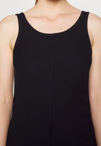 Femme portant une robe noire sans manches avec un col rond, montrée des épaules au milieu du torse sur un fond uni.