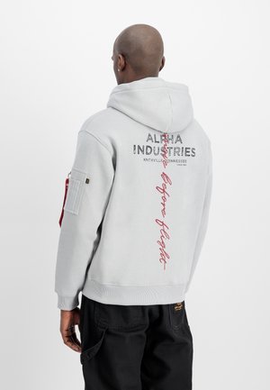 Mężczyzna noszący jasnoszary kaptur Alpha Industries z czarnym i czerwonym napisem na plecach, w połączeniu z czarnymi spodniami, zwrócony tyłem.