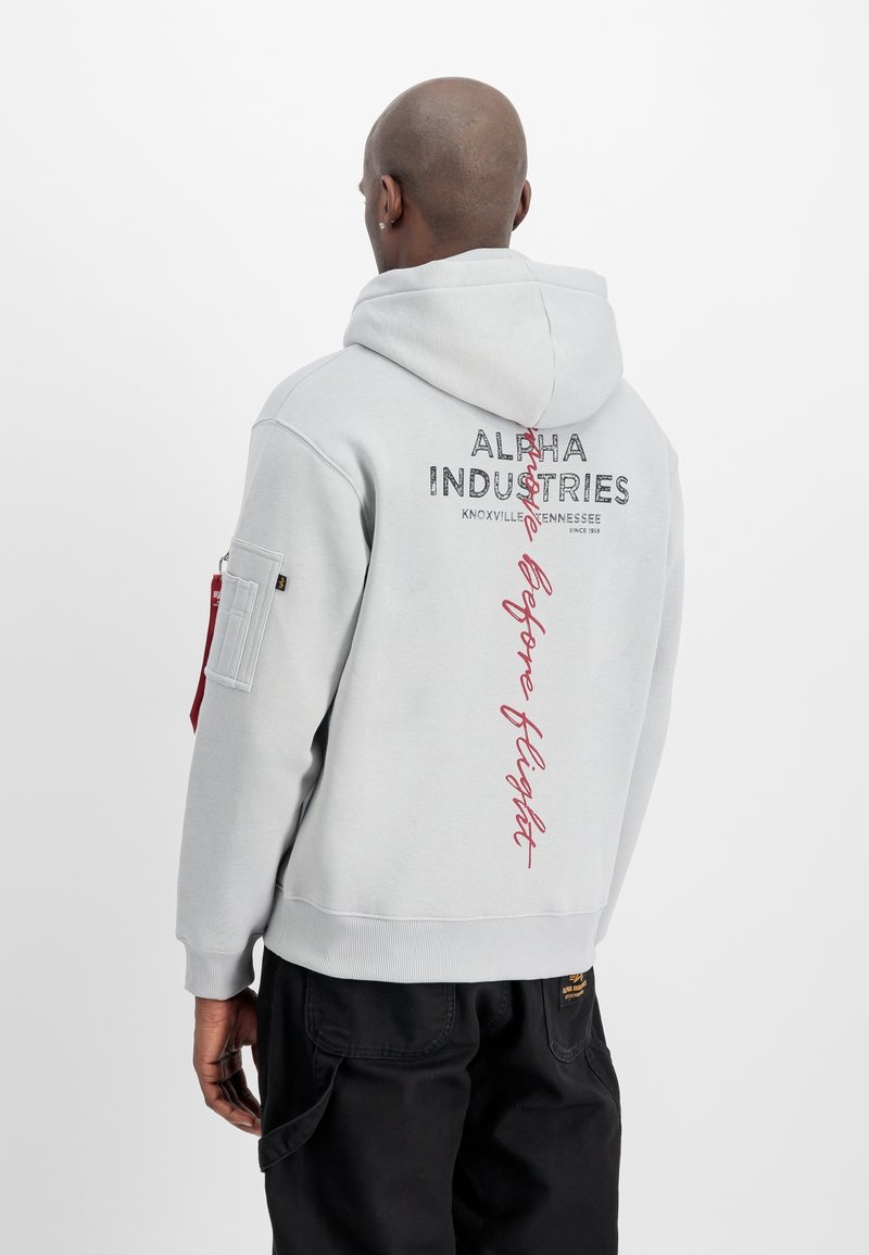 Homme portant un sweat à capuche gris clair Alpha Industries avec un texte noir et rouge au dos, associé à un pantalon noir, de dos.