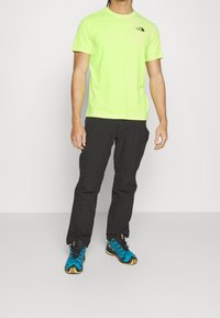 Herren-T-Shirt mit kurzen Ärmeln in Neon-Grün, schwarze Hose und blau-schwarze Outdoorschuhe mit strukturierten Sohlen und strapazierfähigen Materialien.