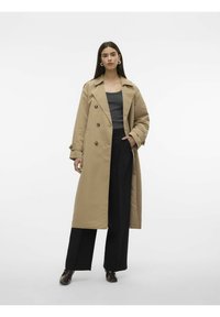Beige dobbeltradet trenchcoat med krage, knapper og lange ermer, båret over en mørk tanktop og svarte vide bukser.