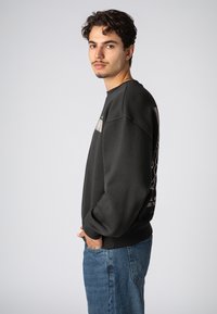 Schwarzer Pullover mit Rundhals, versehen mit einem Logo auf der Brust und einem grafischen Design auf dem Rücken. Getragen mit hellblauen Jeans.
