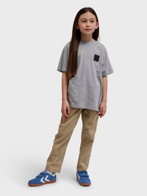 Dziecko o długich włosach, ubrane w szarą koszulkę, beżowe spodnie i niebieskie trampki, stojące na jednolitym jasnym tle.