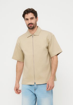 Uomo con capelli scuri e barba che indossa una camicia beige a maniche corte con zip e jeans azzurri, in piedi davanti a uno sfondo chiaro uniforme.