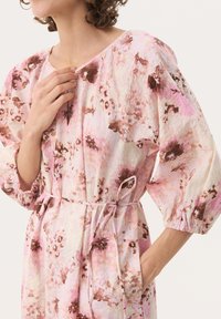 Robe à motifs floraux dans des tons de rose et de beige ; elle présente des manches bouffantes, une taille froncée avec un lien, et des poches latérales. Tissu texturé.