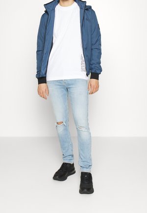 Veste légère bleue avec capuche portée sur un t-shirt blanc, associée à un jean bleu clair déchiré et des baskets noires.