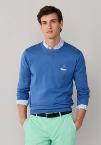 Pull à col rond bleu en tricot doux. Présente un petit motif brodé sur la poitrine et est superposé à une chemise rayée en dessous.