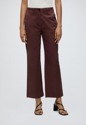 Persoon draagt een bordeauxrode broek met hoge taille en wijde pijpen, een witte top en zwarte sandalen met bandjes en hak, staand tegen een effen achtergrond.