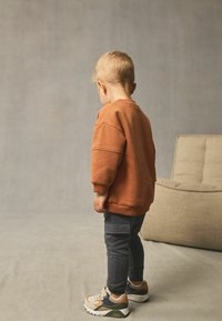 Enfant portant un sweat-shirt orange, un pantalon gris foncé et des baskets beiges avec des accents verts et oranges. Fond minimaliste avec une texture douce.